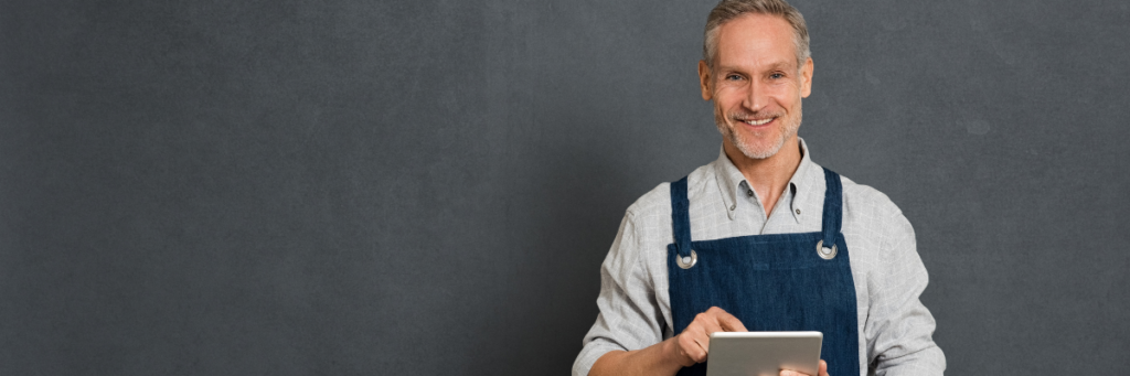 Smiling man standing in front of a dark grey background wearing a blue apron, smiling and using an iPad