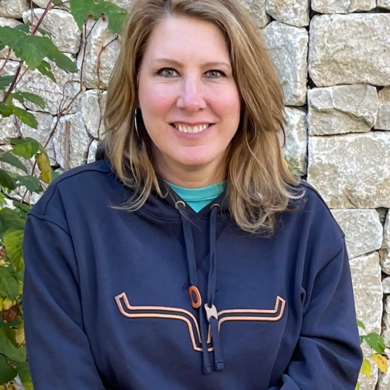 Tonya Day owner of Gather Gift Shop wearing a hoodie and standing outdoors in front of a stone wall