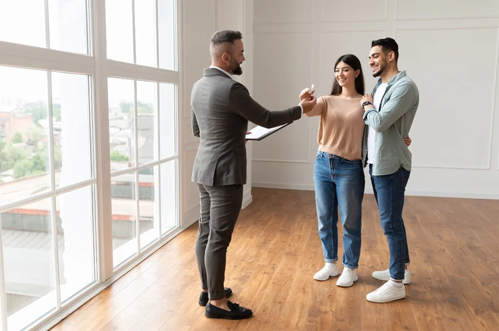 Real estate agent giving keys to a young couple after they have closed on a new home, standing on a wood floor in front of a large window