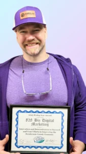 Andrew Murkens, owner of 828 Biz Marketing, standing and smiling in front of a wood background, wearing purple branded clothing and holding a plaque showing that 828 Biz Marketing was named the 2025 Henderson County Chamber of Commerce Rising Star Award Recipient