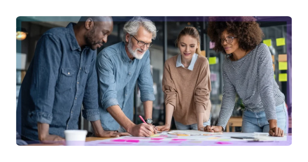 A diverse group of four colleagues collaborate around a table covered with papers and sticky notes, discussing ideas and planning their marketing together in a modern office setting.
