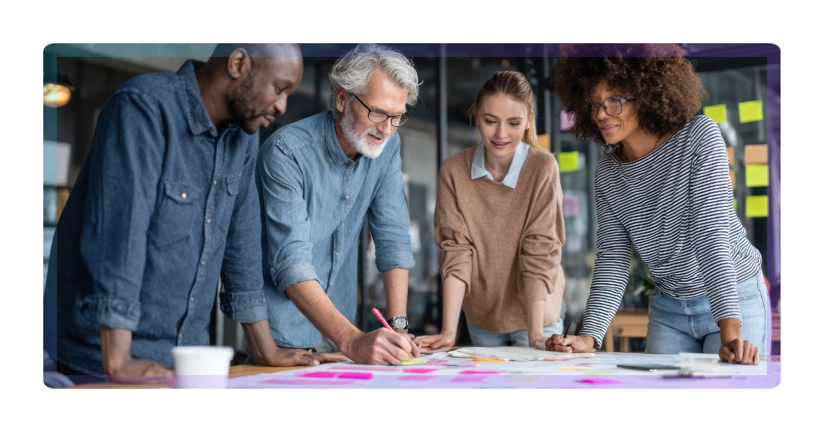 A diverse group of four colleagues collaborate around a table covered with papers and sticky notes, discussing ideas and planning their marketing together in a modern office setting.