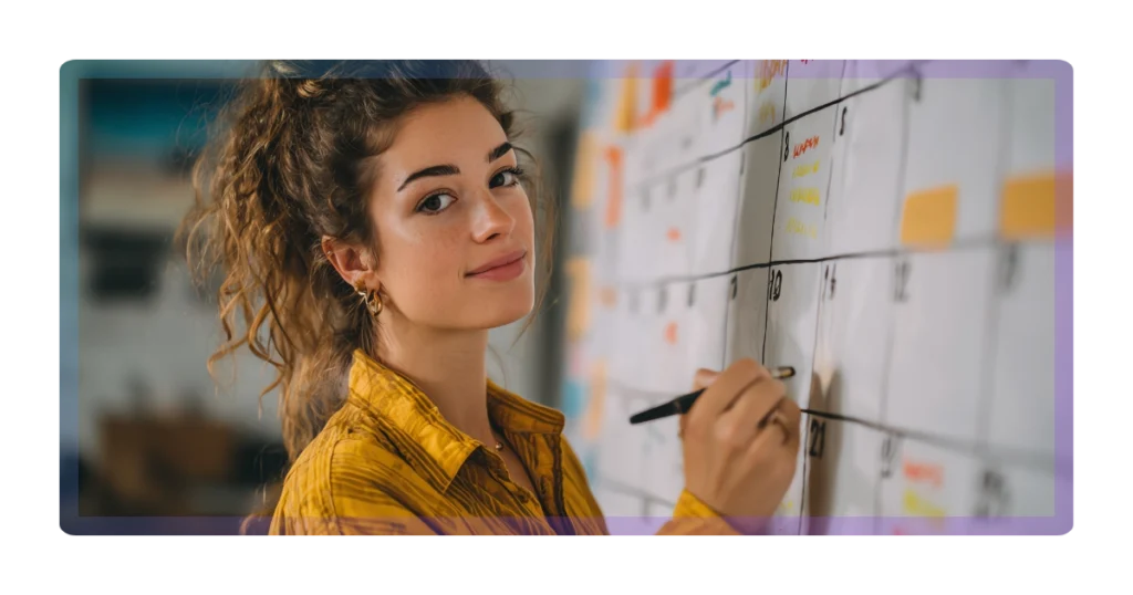 A young woman with curly hair wearing a yellow shirt writes on a large wall calendar with a black marker, planning or organizing her marketing schedule.