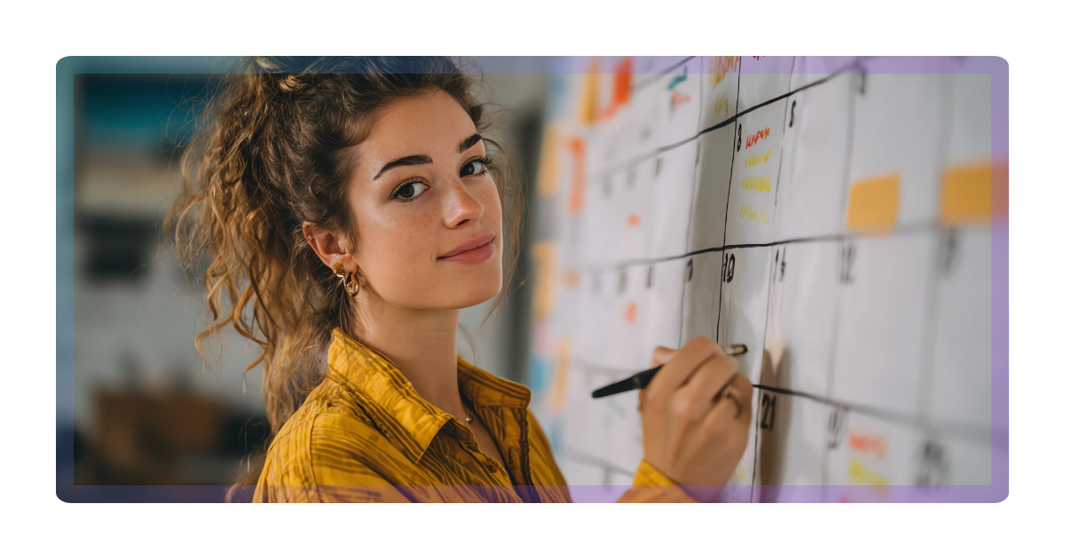 A young woman with curly hair wearing a yellow shirt writes on a large wall calendar with a black marker, planning or organizing her marketing schedule.