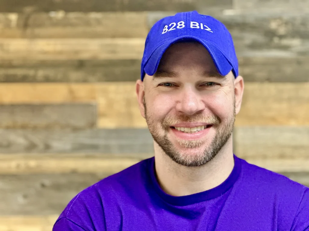 828 Biz Marketing's Founder, Andrew Murkens, smiling, wearing a purple shirt, with a purple 828 Biz branded ball cap, in front of a reclaimed wood background
